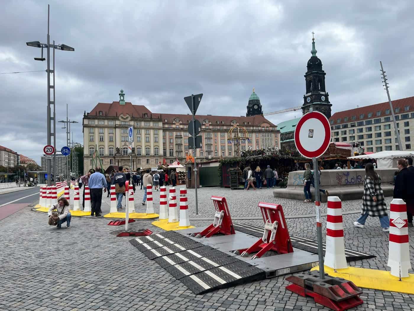 Rot-weiße mobile Zufahrtsperren und Überfahrrampen stehen auf dem Pflaster des Dresdner Altmarkts. Im Hintergrund sind Marktbuden des Herbstmarkts, Passanten sowie die Türme des Rathauses und der Kreuzkirche unter bewölktem Himmel zu sehen.