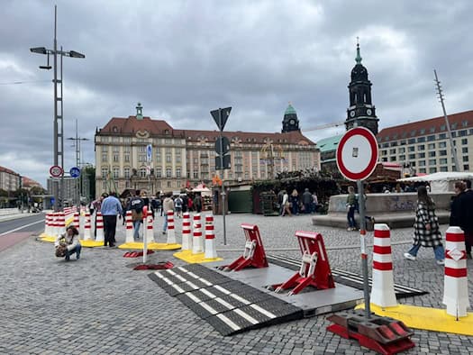 Rot-weiße mobile Zufahrtsperren und Überfahrrampen stehen auf dem Pflaster des Dresdner Altmarkts. Im Hintergrund sind Marktbuden des Herbstmarkts, Passanten sowie die Türme des Rathauses und der Kreuzkirche unter bewölktem Himmel zu sehen.