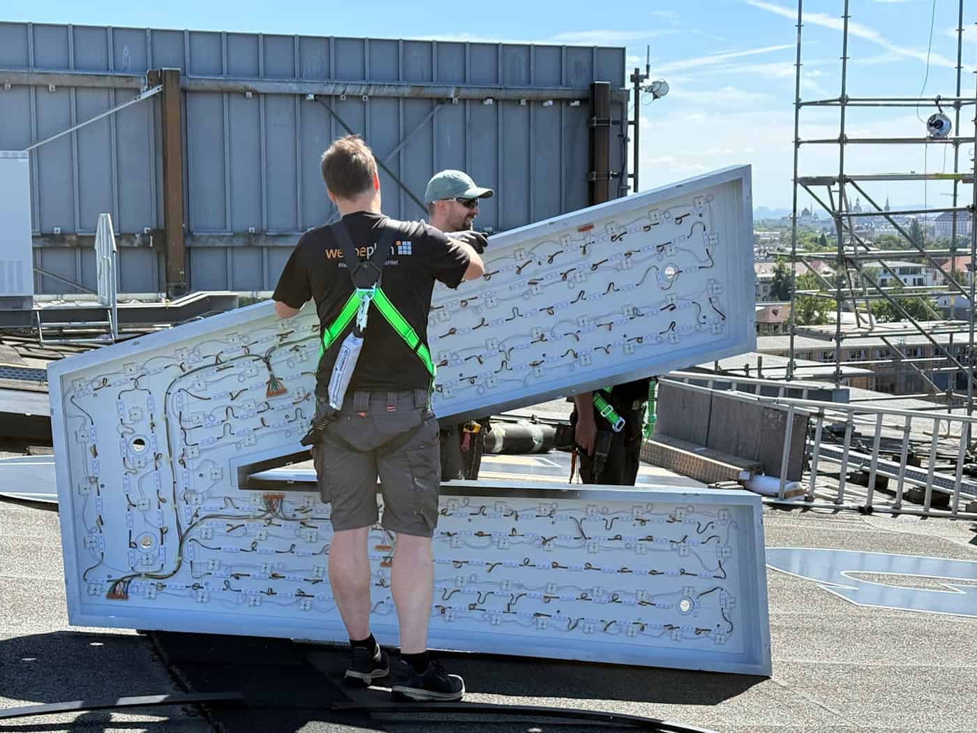 Montage des neuen HIGHVOLT-Schriftzugs auf der Hochspannungshalle Dresden Zwei Handwerker heben auf einem Flachdach das riesige, blau-weiße Buchstabenelement V mit sichtbaren LED-Platinen an, um das neue HIGHVOLT-Logo auf der Hochspannungshalle in Dresden zu montieren.