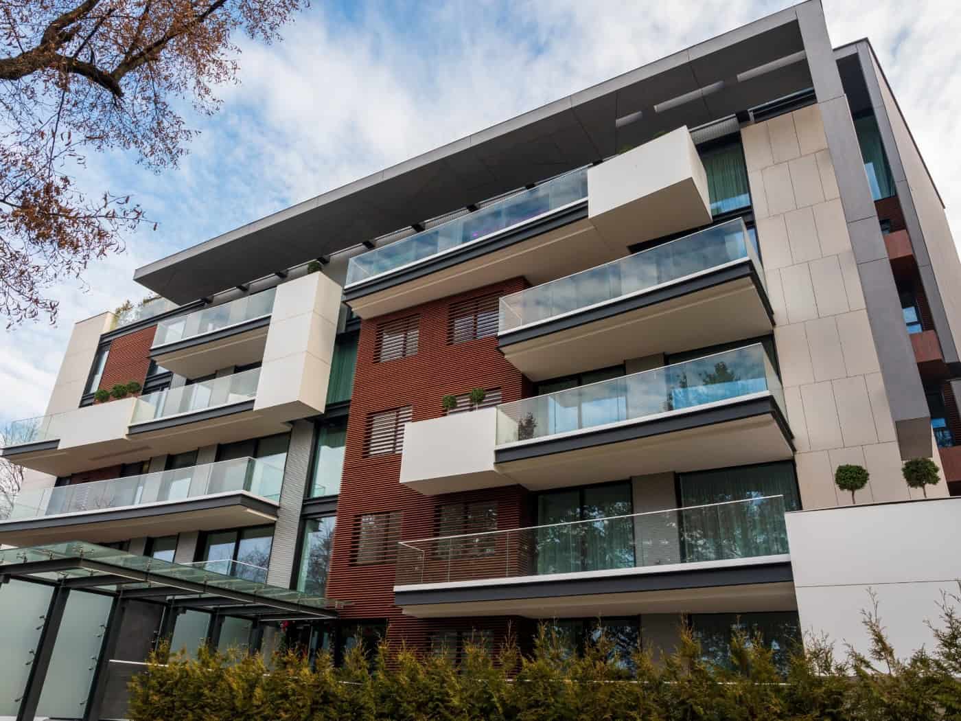 Froschperspektive auf ein modernes, mehrstöckiges Wohnhaus mit Balkonen und Glasgeländern. Die Fassade kombiniert helle Platten mit dunklen Holz- und Metallelementen unter leicht bewölktem blauem Himmel.