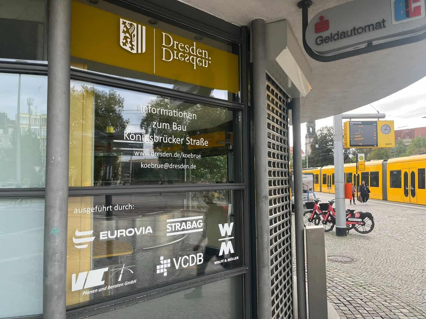 Glaspavillon am Albertplatz mit gelbem Dresden-Logo und der Aufschrift „Informationen zum Bau Königsbrücker Straße“. Rechts im Hintergrund steht eine gelbe Straßenbahn an der DVB-Haltestelle neben einer elektronischen Fahrplananzeige.