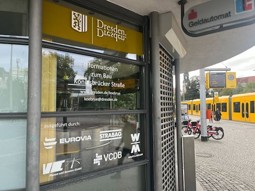Glaspavillon am Albertplatz mit gelbem Dresden-Logo und der Aufschrift „Informationen zum Bau Königsbrücker Straße“. Rechts im Hintergrund steht eine gelbe Straßenbahn an der DVB-Haltestelle neben einer elektronischen Fahrplananzeige.