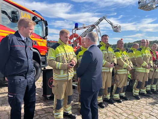 Ein Vertreter der Stadt Dresden im Anzug spricht am Elbufer persönlich mit aufgereihten Feuerwehrleuten in beigefarbener Einsatzkleidung. Dahinter steht ein rotes Tanklöschfahrzeug.   