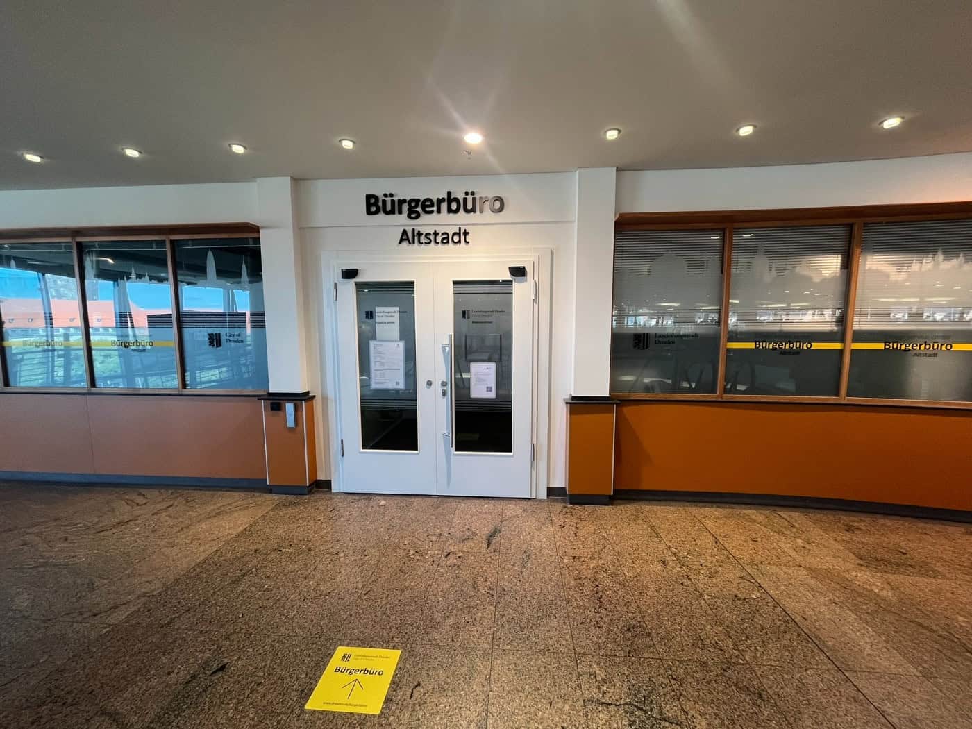 Weiße Eingangstür und Glasfront des Bürgerbüros Altstadt im Kaufhaus Galeria mit Beschriftung über der Tür und gelbem Richtungspfeil auf dem Steinboden vor dem Eingang.