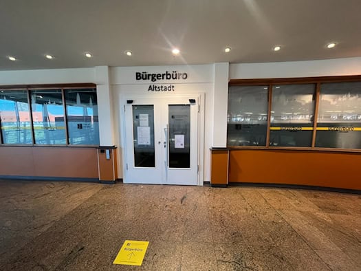 Weiße Eingangstür und Glasfront des Bürgerbüros Altstadt im Kaufhaus Galeria mit Beschriftung über der Tür und gelbem Richtungspfeil auf dem Steinboden vor dem Eingang.