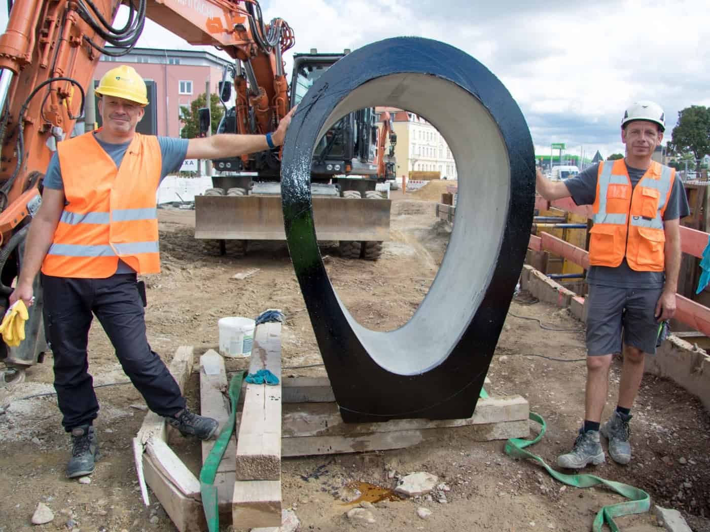 Zwei Bauarbeiter mit Helmen und Warnwesten stehen links und rechts neben einem großen, hochkant aufgestellten Beton-Eiprofil für den Kanalbau auf einer Erdbaustelle. Im Hintergrund steht ein orangefarbener Kettenbagger.