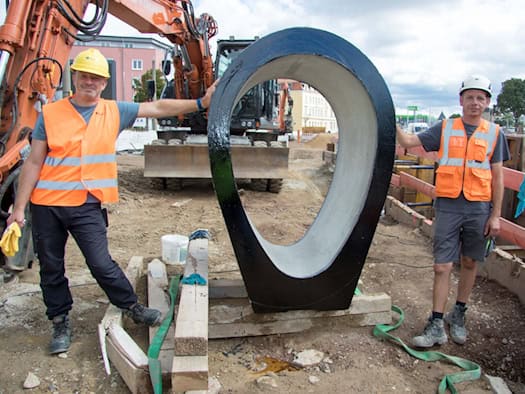 Zwei Bauarbeiter mit Helmen und Warnwesten stehen links und rechts neben einem großen, hochkant aufgestellten Beton-Eiprofil für den Kanalbau auf einer Erdbaustelle. Im Hintergrund steht ein orangefarbener Kettenbagger.