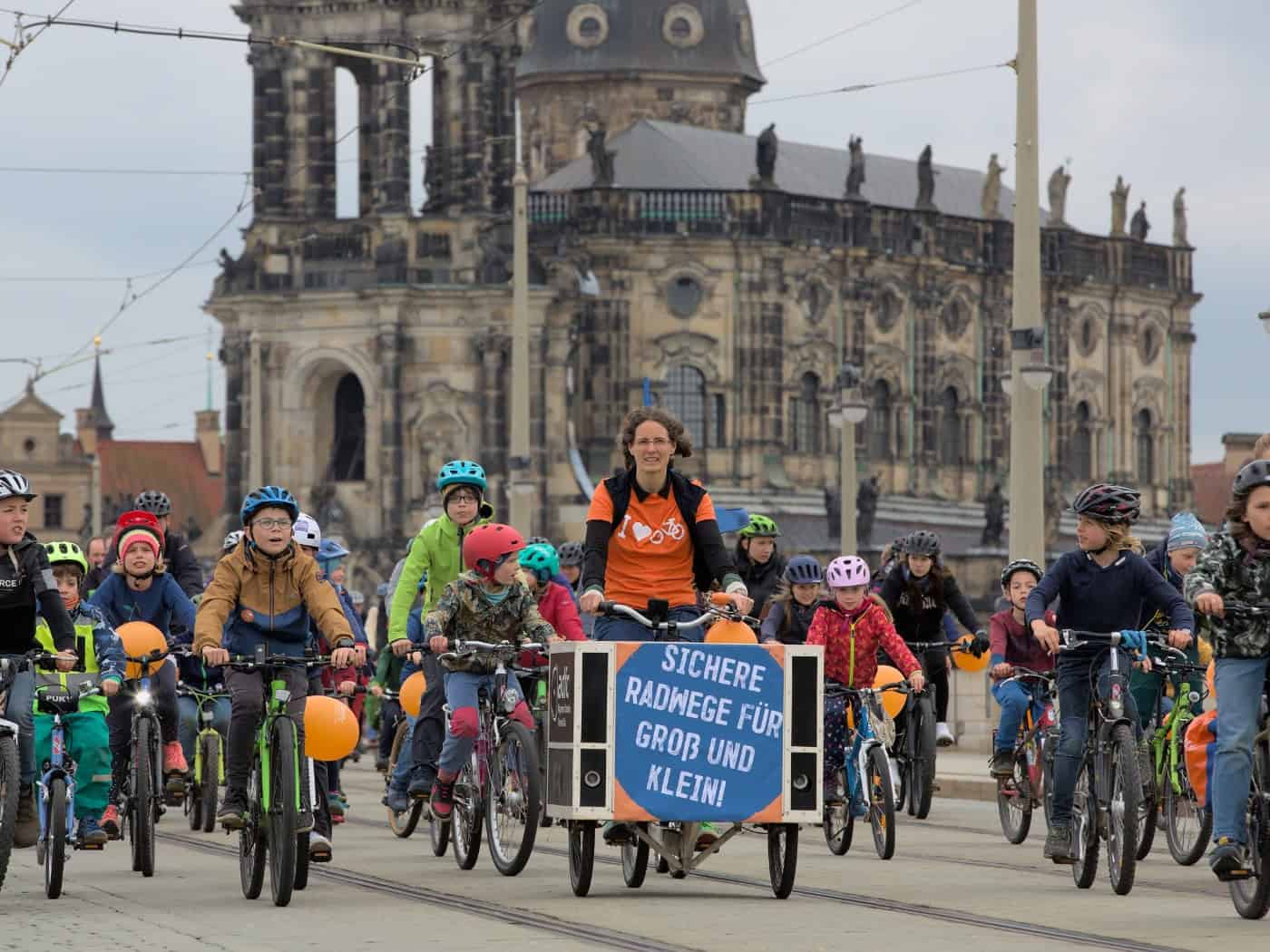 Eine Gruppe von Kindern und Erwachsenen fährt auf Fahrrädern über eine Straße in Dresden. Eine Frau steuert ein Lastenrad mit dem Schild „Sichere Radwege für Groß und Klein!“. Im Hintergrund ist die Katholische Hofkirche vor leicht bewölktem Himmel zu sehen.