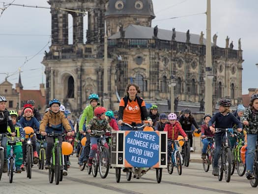 Eine Gruppe von Kindern und Erwachsenen fährt auf Fahrrädern über eine Straße in Dresden. Eine Frau steuert ein Lastenrad mit dem Schild „Sichere Radwege für Groß und Klein!“. Im Hintergrund ist die Katholische Hofkirche vor leicht bewölktem Himmel zu sehen.