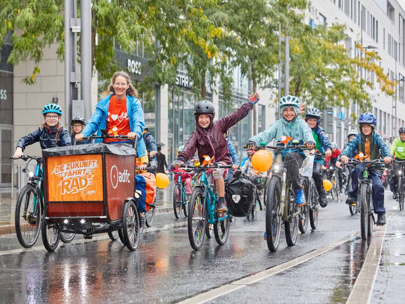 Eine Gruppe fröhlicher Kinder und Erwachsener radelt mit Fahrradhelmen über eine nassgeregnete Straße in der Stadt. Mittig fährt ein Lastenfahrrad mit einem orangefarbenen ADFC-Schild mit der Aufschrift „Die Zukunft fährt Rad“. Die Kinder lächeln und winken.