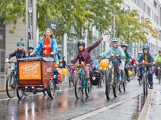 Eine Gruppe fröhlicher Kinder und Erwachsener radelt mit Fahrradhelmen über eine nassgeregnete Straße in der Stadt. Mittig fährt ein Lastenfahrrad mit einem orangefarbenen ADFC-Schild mit der Aufschrift „Die Zukunft fährt Rad“. Die Kinder lächeln und winken.