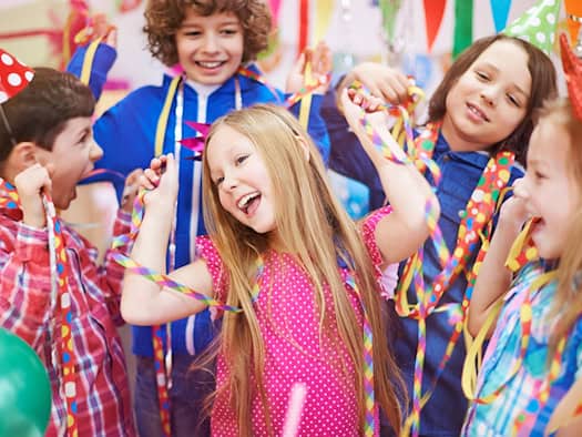 Eine Gruppe fröhlicher Kinder feiert ausgelassen mit bunter Partydekoration. Sie tragen bunte Spitzhüte, tanzen und halten bunte Luftschlangen sowie Wimpelketten in den Händen. Die Stimmung ist ausgelassen und festlich.