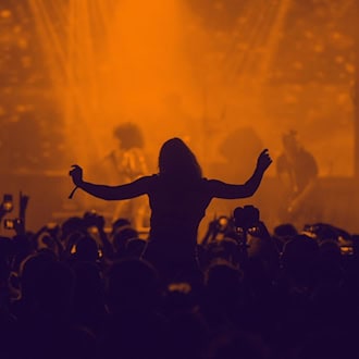 Silhouette einer feiernden Menschenmenge bei einem Konzert im warmen, orangefarbenen Bühnenlicht. In der Mitte tanzt eine Person auf den Schultern einer anderen mit erhobenen Armen, während Besucher das Geschehen mit Smartphones festhalten.