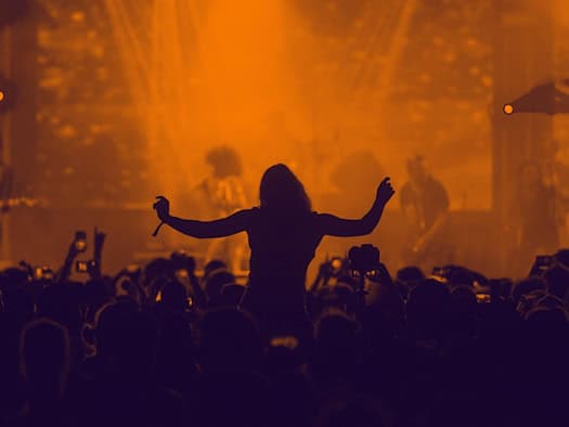 Silhouette einer feiernden Menschenmenge bei einem Konzert im warmen, orangefarbenen Bühnenlicht. In der Mitte tanzt eine Person auf den Schultern einer anderen mit erhobenen Armen, während Besucher das Geschehen mit Smartphones festhalten.