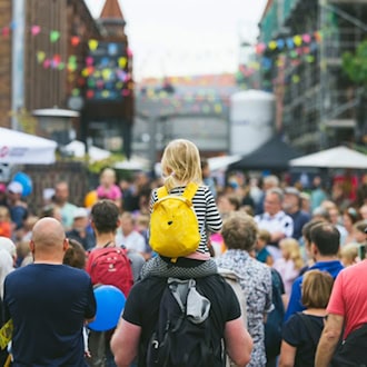 Blick von hinten auf eine dichte Menschenmenge bei einem Sommerfest. In der Mitte trägt ein Mann ein kleines Kind mit einem gelben Tier-Rucksack auf den Schultern. Im Hintergrund stehen Backsteingebäude des Kraftwerks Mitte mit bunten Wimpelketten.