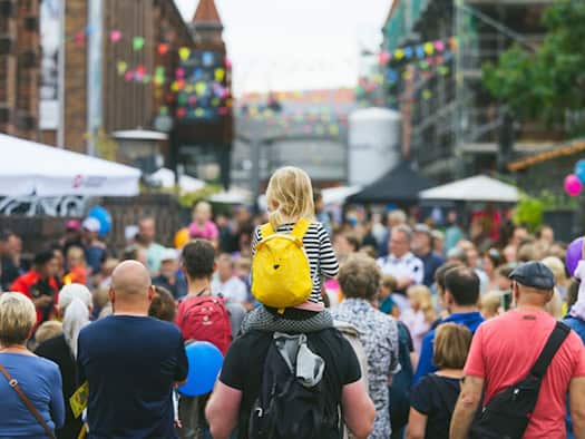 Blick von hinten auf eine dichte Menschenmenge bei einem Sommerfest. In der Mitte trägt ein Mann ein kleines Kind mit einem gelben Tier-Rucksack auf den Schultern. Im Hintergrund stehen Backsteingebäude des Kraftwerks Mitte mit bunten Wimpelketten.