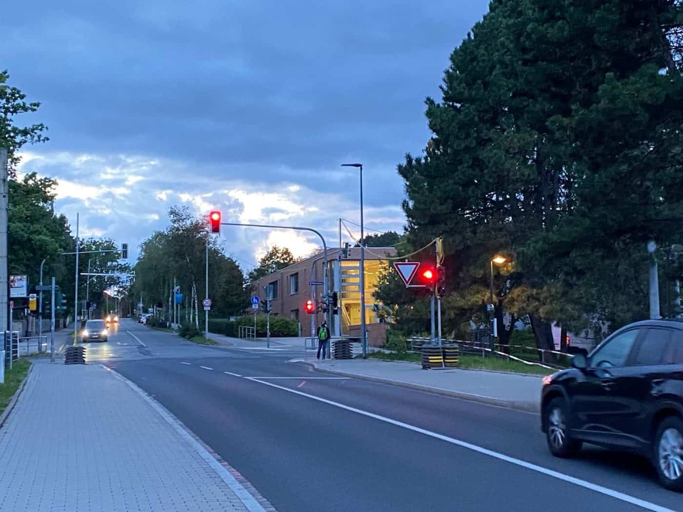 Blick bei Abenddämmerung auf eine asphaltierte Straße mit roter Ampel in Dresden-Räcknitz. Links verläuft ein gepflasterter Gehweg, rechts ist ein schwarzes Auto zu sehen. Im Hintergrund steht ein beleuchtetes Gebäude neben Bäumen unter dämmrigem Himmel.