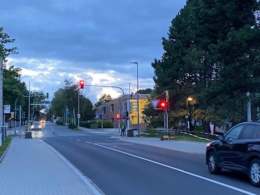 Blick bei Abenddämmerung auf eine asphaltierte Straße mit roter Ampel in Dresden-Räcknitz. Links verläuft ein gepflasterter Gehweg, rechts ist ein schwarzes Auto zu sehen. Im Hintergrund steht ein beleuchtetes Gebäude neben Bäumen unter dämmrigem Himmel.