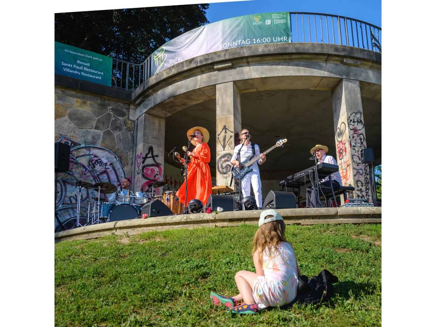 Blick von einer Wiese auf einen Steinpavillon in Dresden: Eine Band mit Sängern, Bassist, Drummer und Keyboarder tritt auf der überdachten Bühne auf. Im Vordergrund sitzt ein kleines Mädchen im Gras und schaut zu.