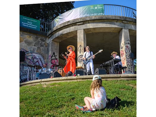 Blick von einer Wiese auf einen Steinpavillon in Dresden: Eine Band mit Sängern, Bassist, Drummer und Keyboarder tritt auf der überdachten Bühne auf. Im Vordergrund sitzt ein kleines Mädchen im Gras und schaut zu.