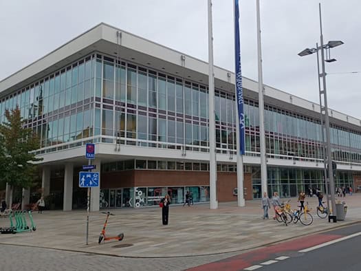Außenansicht des Dresdner Kulturpalasts mit moderner Glasfassade und Flachdach. Auf dem großen Vorplatz stehen Bänke, Fahrräder und E-Scooter, während einige Passanten vorbeigehen.   