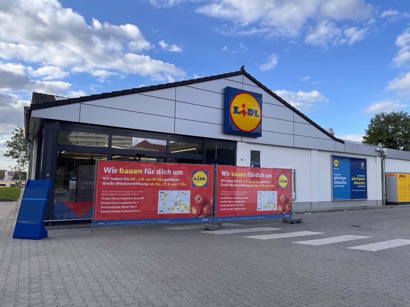 Außenansicht eines Lidl-Supermarktes bei leicht bewölktem Himmel. Im Vordergrund stehen zwei rote Werbebanner mit der Aufschrift „Wir bauen für dich um“ und Hinweisen zur Wiedereröffnung vor dem Eingangsbereich auf dem gepflasterten Parkplatz.