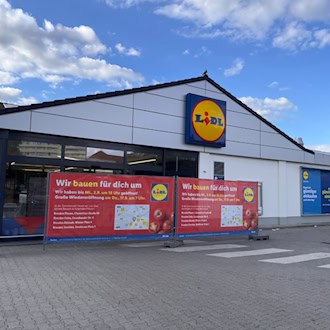 Außenansicht eines Lidl-Supermarktes bei leicht bewölktem Himmel. Im Vordergrund stehen zwei rote Werbebanner mit der Aufschrift „Wir bauen für dich um“ und Hinweisen zur Wiedereröffnung vor dem Eingangsbereich auf dem gepflasterten Parkplatz.