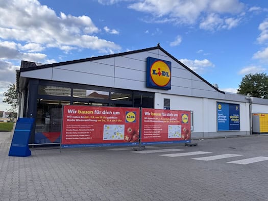 Außenansicht eines Lidl-Supermarktes bei leicht bewölktem Himmel. Im Vordergrund stehen zwei rote Werbebanner mit der Aufschrift „Wir bauen für dich um“ und Hinweisen zur Wiedereröffnung vor dem Eingangsbereich auf dem gepflasterten Parkplatz.