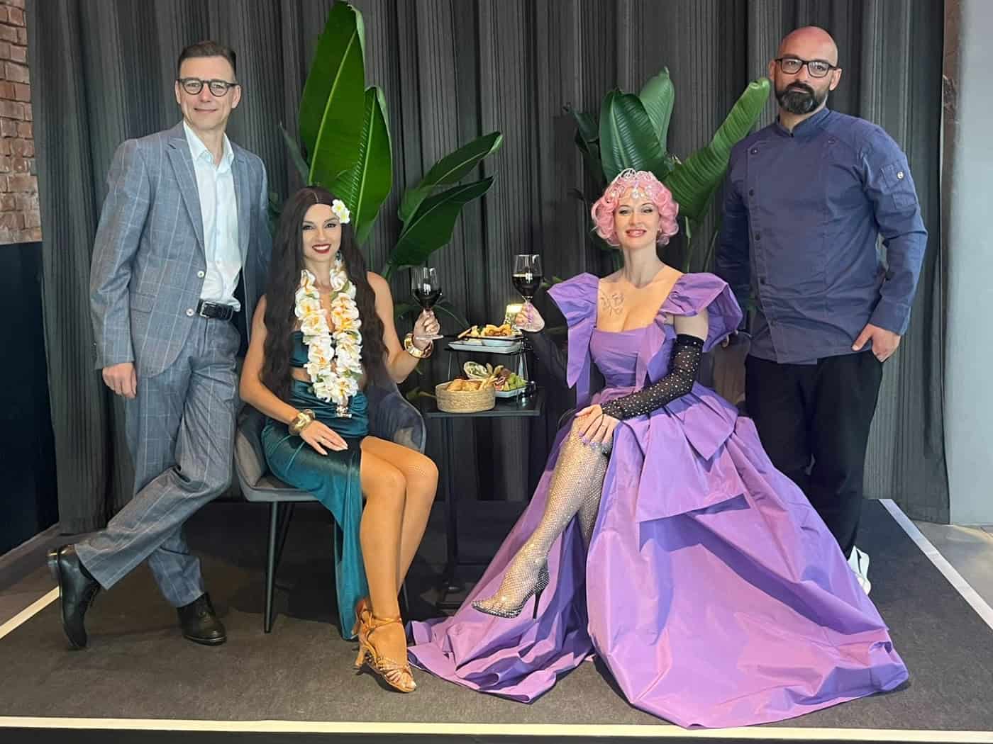 Auf einem kleinen Podest posieren vier Personen: Guido Gentzel im Anzug, eine Frau im grünen Kleid mit Blumenkette, eine Frau im üppigen violetten Abendkleid und Koch Sven Kasimir in dunkler Kochjacke. Im Hintergrund stehen Zimmerpflanzen vor einem dunklen Vorhang.