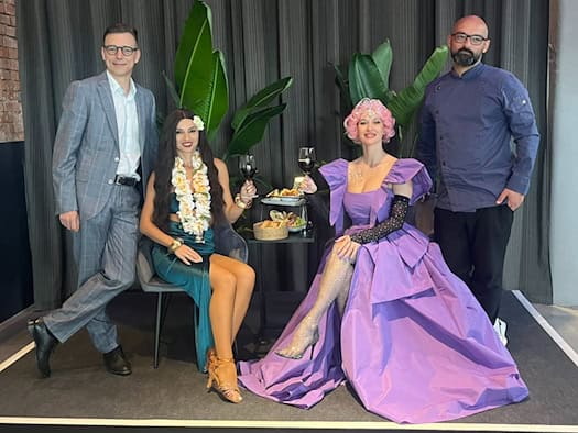 Auf einem kleinen Podest posieren vier Personen: Guido Gentzel im Anzug, eine Frau im grünen Kleid mit Blumenkette, eine Frau im üppigen violetten Abendkleid und Koch Sven Kasimir in dunkler Kochjacke. Im Hintergrund stehen Zimmerpflanzen vor einem dunklen Vorhang.