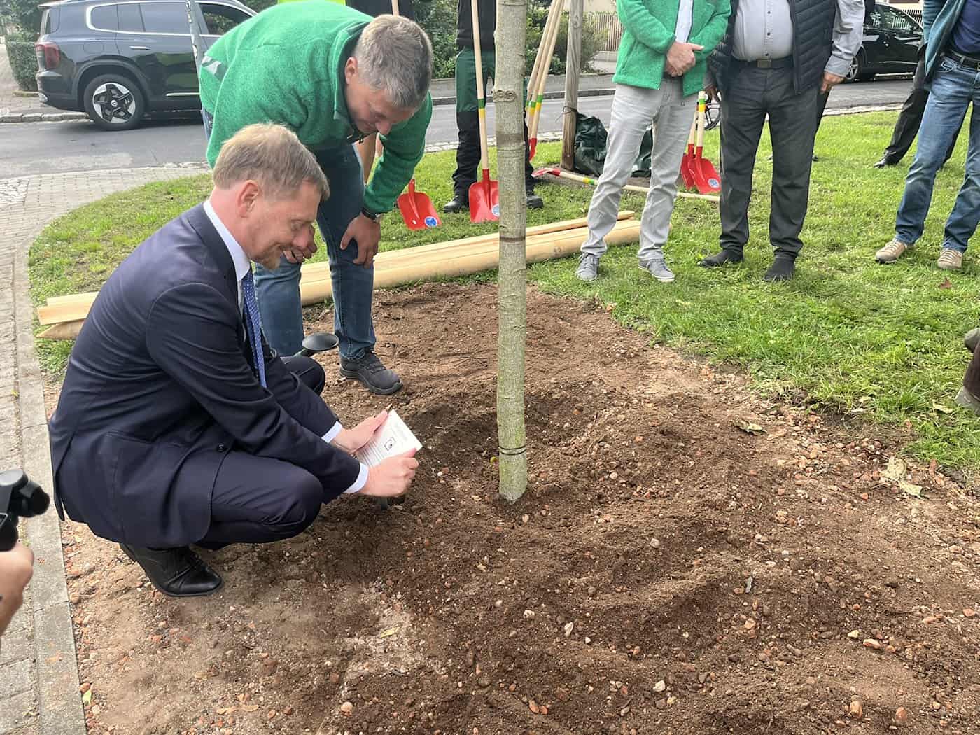 Nahaufnahme von Sachsens Ministerpräsident Michael Kretschmer, der im dunklen Anzug am Boden neben dem Stamm eines jung gepflanzten Baumes hockt. Ein Gärtner in grüner Arbeitskleidung steht gebeugt direkt hinter ihm auf der frischen Erde.