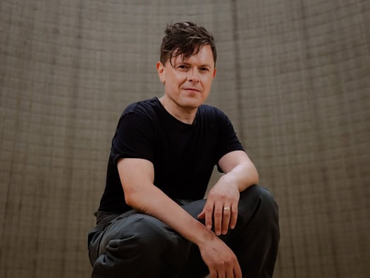 Porträt von Sänger Michael Patrick Kelly in dunklem T-Shirt, wie er leicht hockend direkt in die Kamera blickt, vor einem strukturierten, neutralen Hintergrund.  