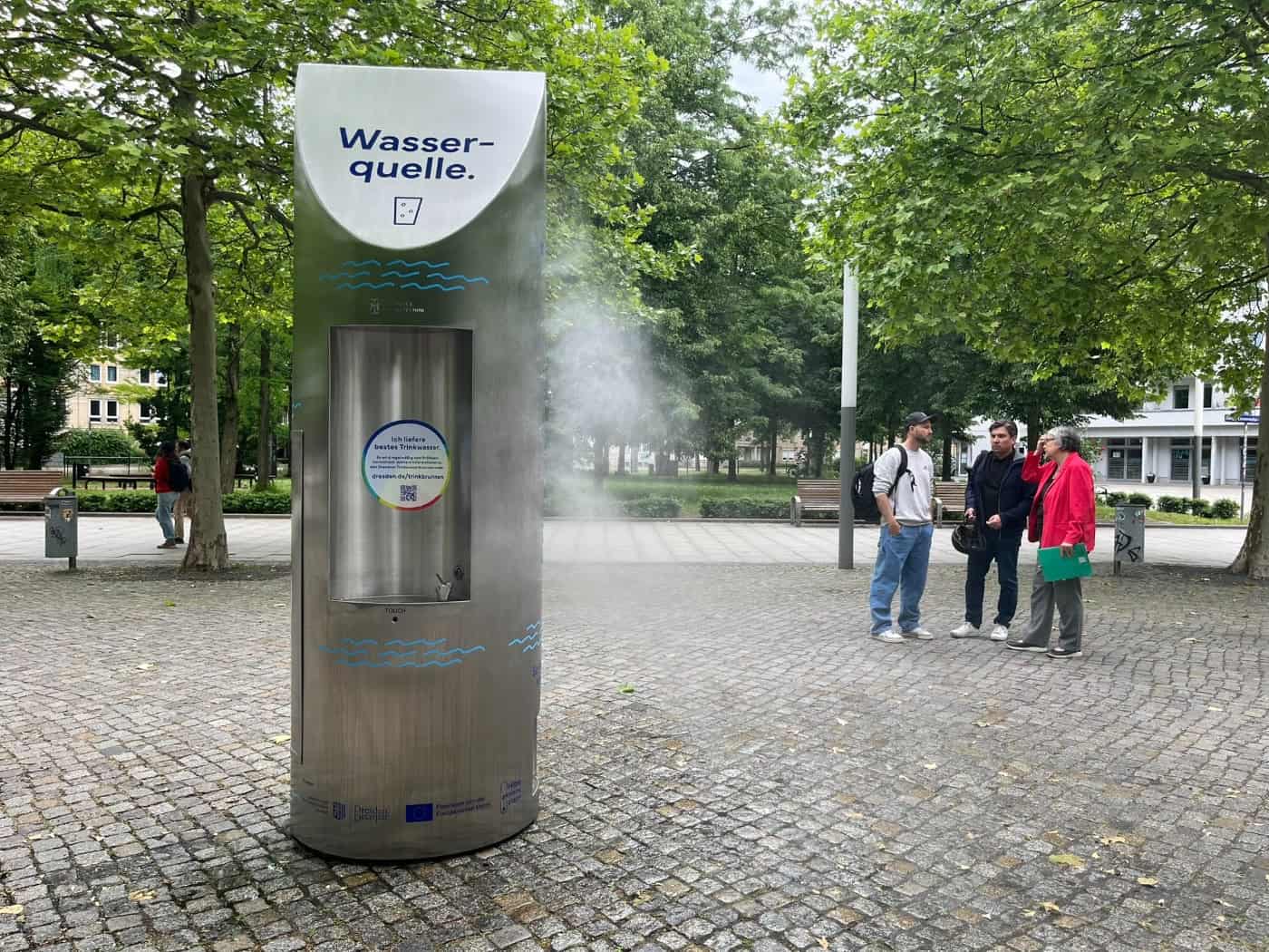Eine silberne Metall-Trinkwasserstele mit der Aufschrift „Wasserquelle“ versprüht feinen Feuchtigkeitsnebel auf einem gepflasterten Platz. Drei Personen beobachten die Anlage, im Hintergrund stehen grüne Bäume.