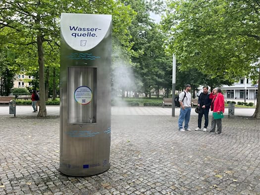 Eine silberne Metall-Trinkwasserstele mit der Aufschrift „Wasserquelle“ versprüht feinen Feuchtigkeitsnebel auf einem gepflasterten Platz. Drei Personen beobachten die Anlage, im Hintergrund stehen grüne Bäume.