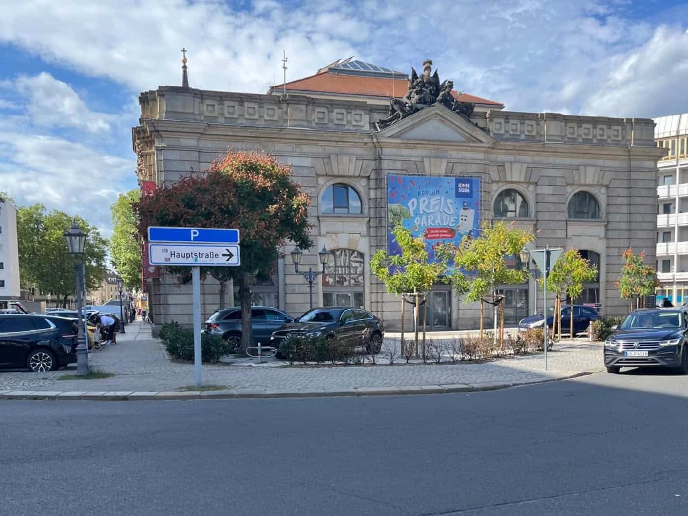 Außenansicht der historischen Neustädter Markthalle aus Sandstein in Dresden unter leicht bewölktem Himmel. Im Vordergrund verläuft eine Straße mit Parkschild („Hauptstraße“), davor stehen Bäume und geparkte Autos.