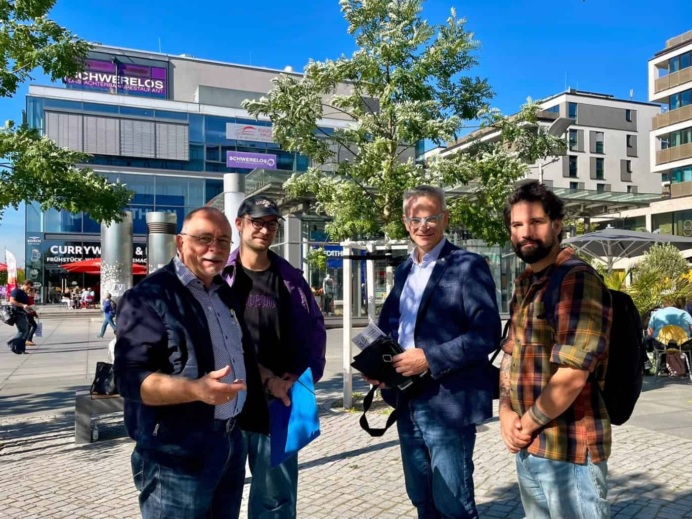 Vier Männer stehen draußen auf dem sonnigen Wiener Platz in Dresden: André Barth, Peter Henker mit Mütze, MdB Markus Reichel im Sakko und Sozialarbeiter Alexander Beuschel mit Rucksack (v.l.n.r.). Im Hintergrund ein Geschäftsgebäude.