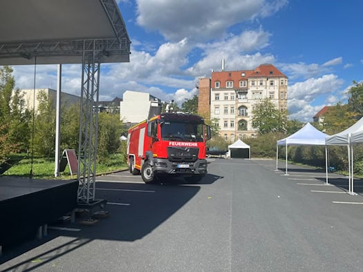 Ein rotes Feuerwehrfahrzeug parkt auf einem Teerplatz zwischen einer schwarzen Veranstaltungsbühne links und weißen Zelten rechts. Im Hintergrund steht ein helles Gründerzeitgebäude unter leicht bewölktem blauem Himmel.