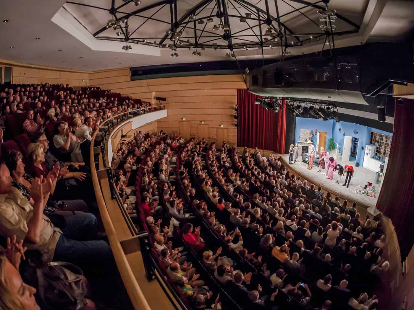 Weitwinkelaufnahme des gefüllten Theatersaals der Comödie Dresden von oben. Das Publikum auf den Rängen und im Parkett klatscht Beifall, während auf der beleuchteten Bühne die Schauspieler in Kostümen stehen und sich vor dem roten Vorhang verbeugen.
