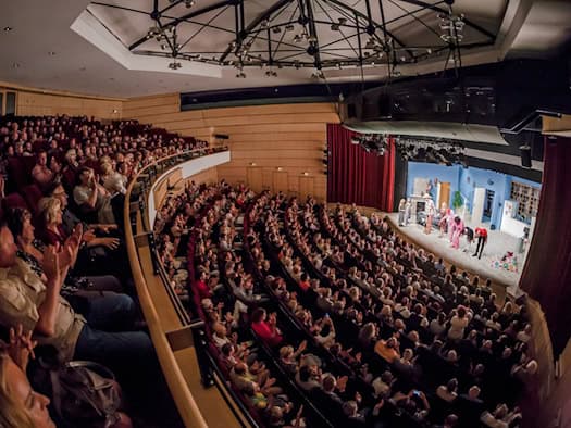 Weitwinkelaufnahme des gefüllten Theatersaals der Comödie Dresden von oben. Das Publikum auf den Rängen und im Parkett klatscht Beifall, während auf der beleuchteten Bühne die Schauspieler in Kostümen stehen und sich vor dem roten Vorhang verbeugen.