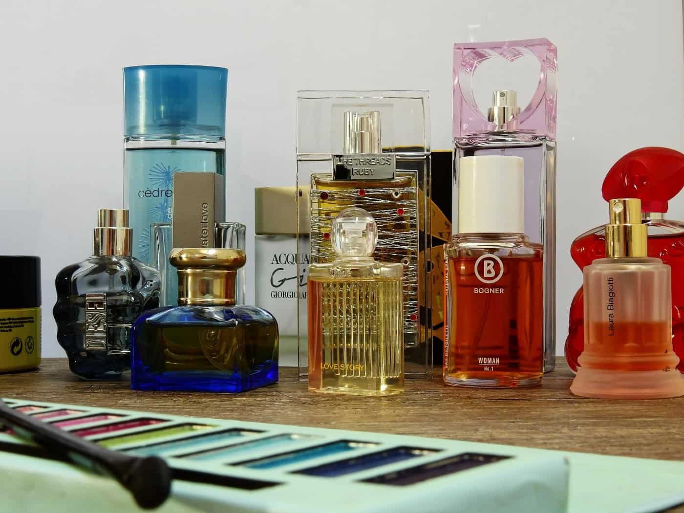Symbolbild zum Parfümdiebstahl: Verschiedene Parfümflakons in unterschiedlichen Formen und Farben stehen auf einer Holzoberfläche. Im Vordergrund liegt eine geöffnete Lidschattenpalette mit einem Kosmetikpinsel.