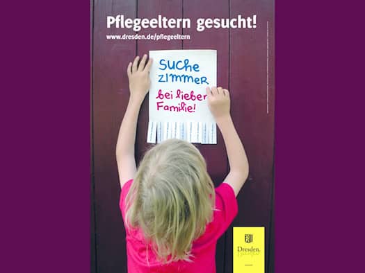 braunen Holzwand. Auf dem Zettel steht in Buntstift-Optik „Suche Zimmer bei lieber Familie!“, darüber der Titel „Pflegeeltern gesucht!“.