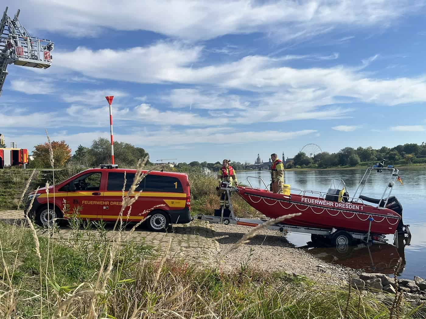 Ein roter Mannschaftswagen der Feuerwehr bringt ein rotes Rettungsboot auf einem Trailer über die gepflasterte Slipstelle direkt ins Wasser der Elbe. Zwei Feuerwehrmänner stehen im Boot.
