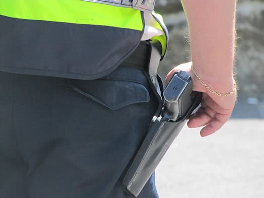 Nahaufnahme der Hand eines Polizisten an seiner Dienstwaffe, die in einem schwarzen Lederholster am Gürtel steckt. Der Beamte trägt eine gelbe Warnweste und eine dunkle Hose.