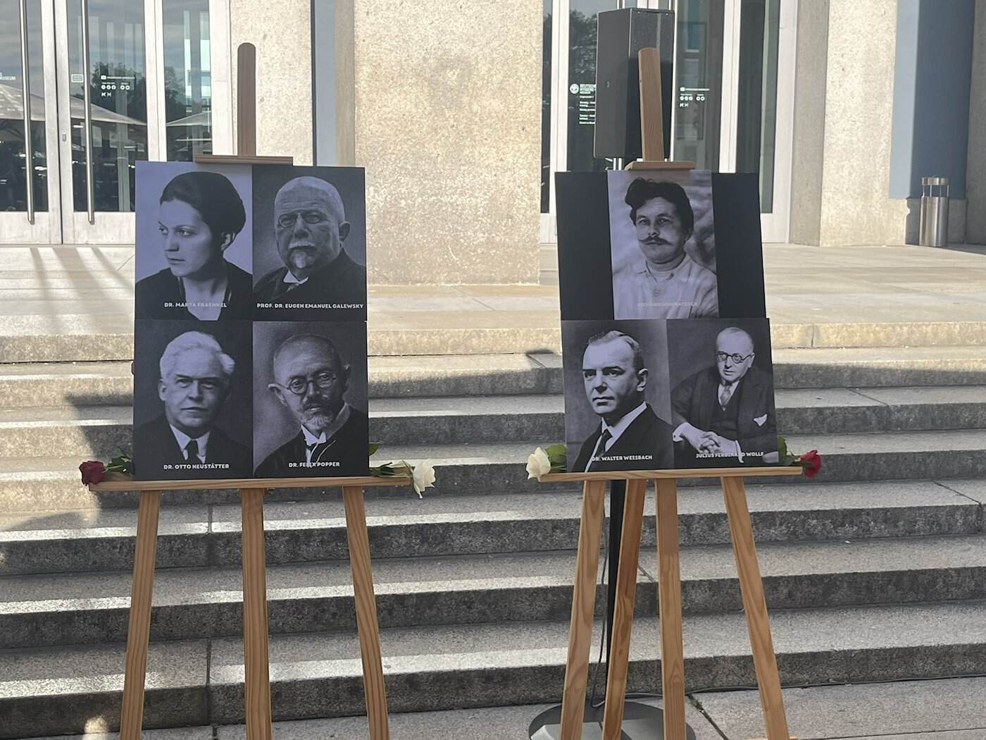 Zwei Holzstaffeleien stehen auf den Stufen vor dem Eingang des Museums. Darauf sind historische Schwarz-Weiß-Porträts der sieben Menschen angebracht, an die mit Stolpersteinen erinnert wird. Kleine Rosen liegen davor.