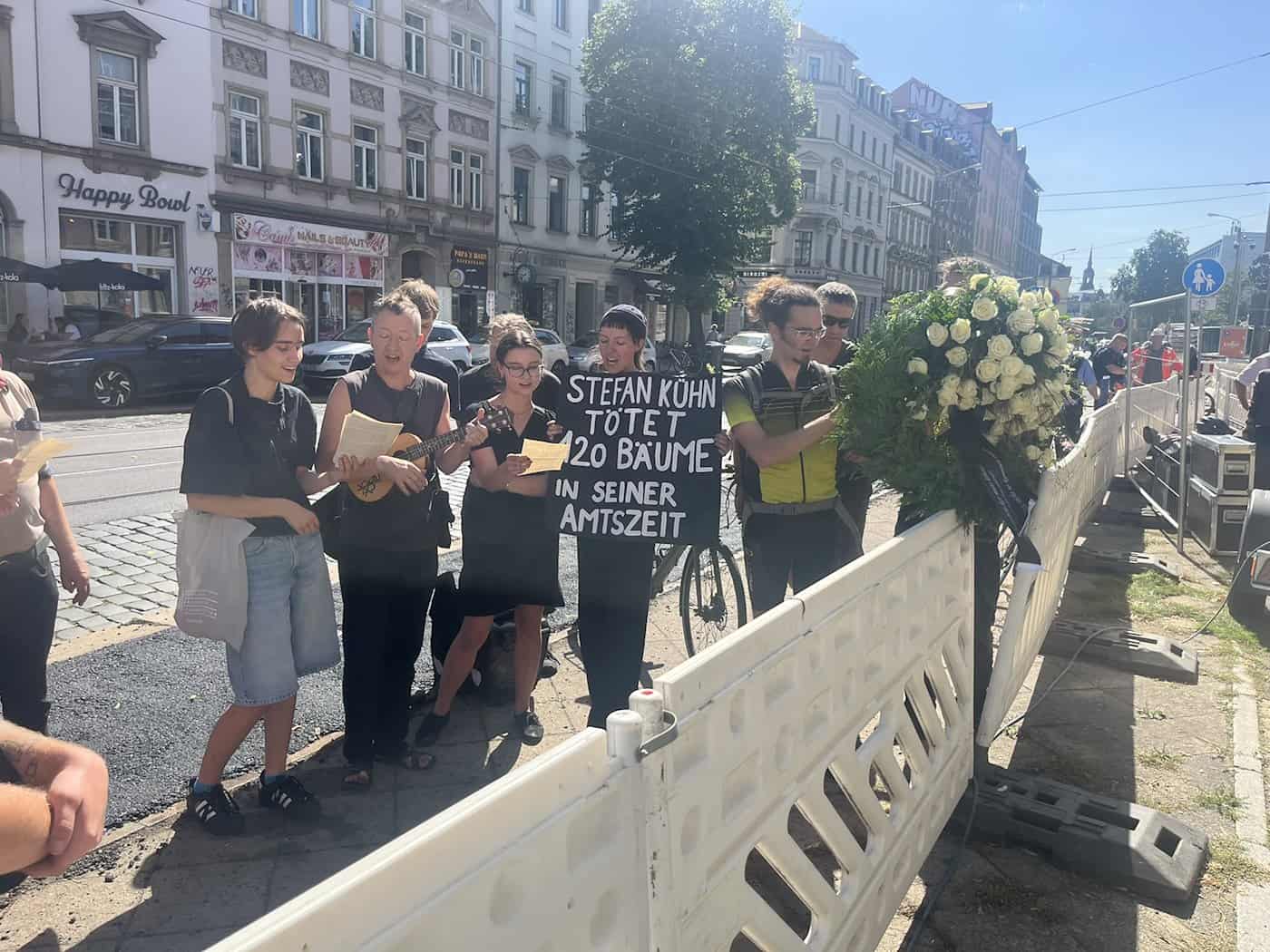 Eine Gruppe von Aktivisten steht hinter einem weißen Bauzaun auf dem Gehweg. Eine Person spielt Ukulele, eine andere hält ein Pappschild mit der Aufschrift „Stefan Kühn tötet 120 Bäume in seiner Amtszeit“. Neben ihnen hängt ein großer Trauerkranz.