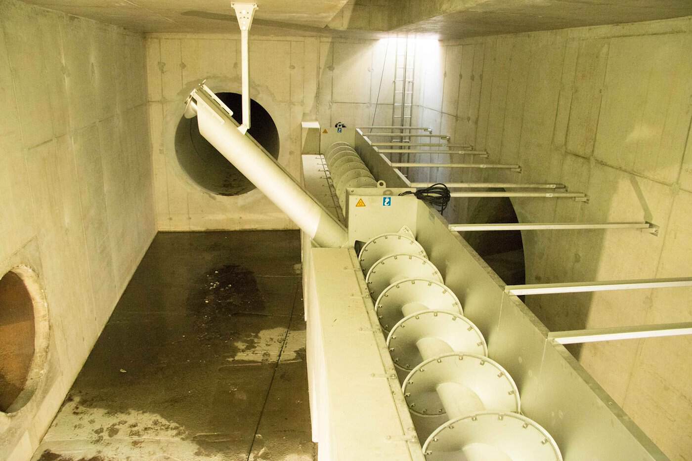 Ein unterirdischer, quaderförmiger Betonraum mit großen Rohreingängen in den Wänden. In der Mitte verläuft eine mechanische Förderschnecke aus Metall zur Reinigung von Abwasser.