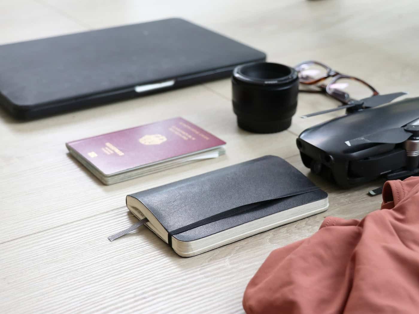 Auf hellem Holzfußboden liegen ein roter deutscher Reisepass, ein schwarzes Notizbuch, ein Laptop, eine Brille, ein Kameraobjektiv und eine kleine Drohne.