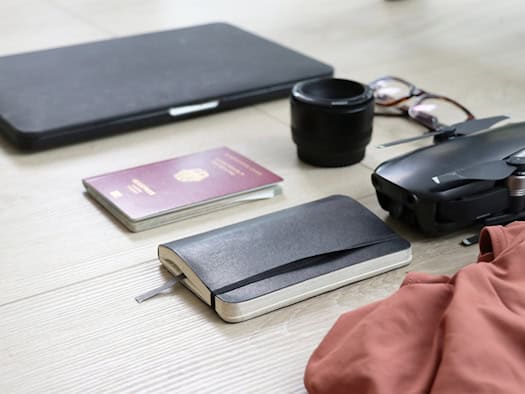 Auf hellem Holzfußboden liegen ein roter deutscher Reisepass, ein schwarzes Notizbuch, ein Laptop, eine Brille, ein Kameraobjektiv und eine kleine Drohne.