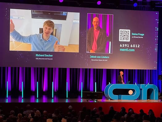 Scénická obrazovka na konferenci o umělé inteligenci v Drážďanech: Na velkém plátně je vidět připojený zakladatel Richard Socher v světle modré košili. Vpravo vedle něj se objevuje moderátor Jakob von Lindern. Scénu osvětlují fialová světla.