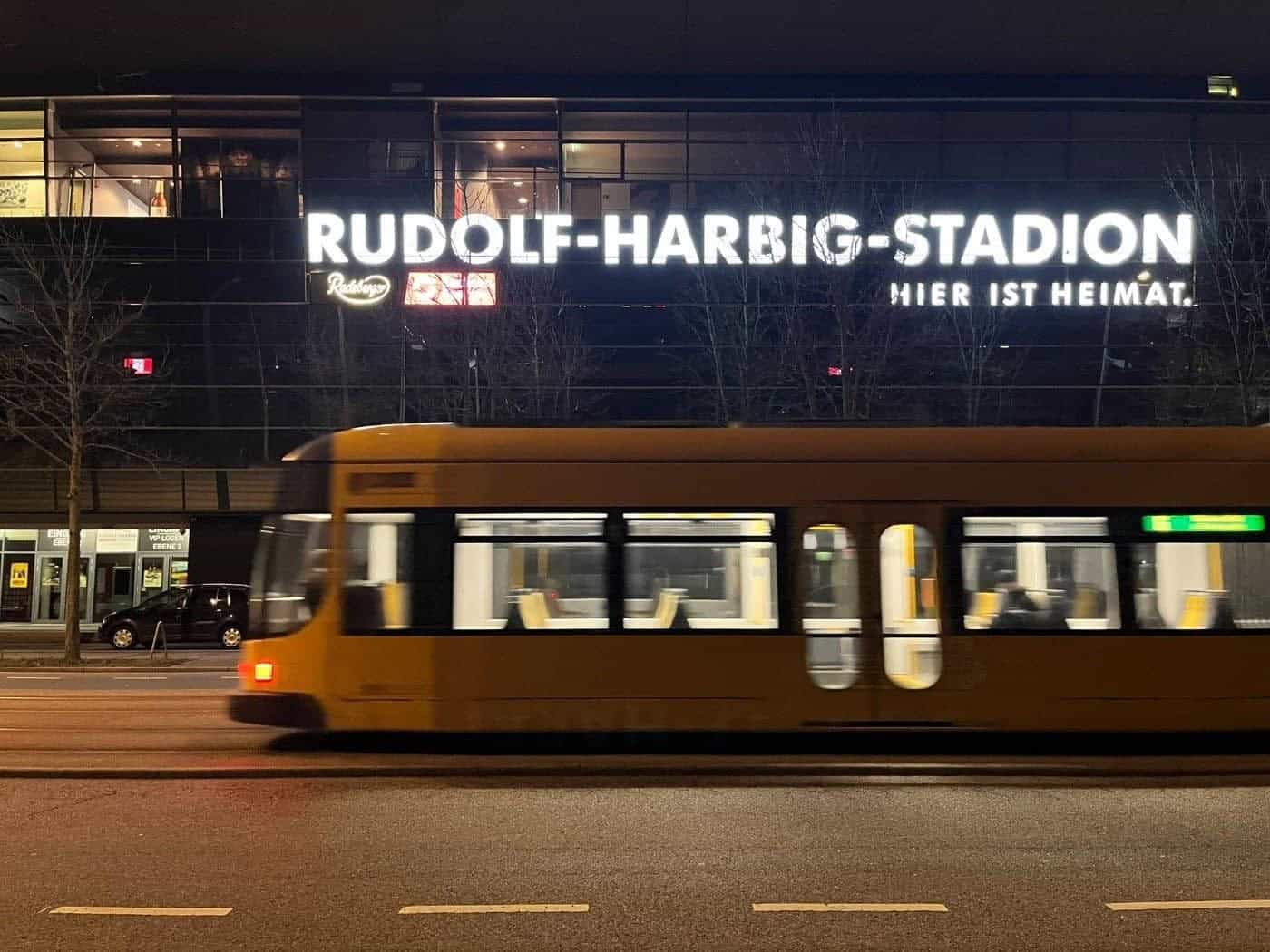 Nachtaufnahme des Rudolf-Harbig-Stadions in Dresden mit leuchtender Schrift „Rudolf-Harbig-Stadion Hier ist Heimat“. Im Vordergrund fährt eine gelbe DVB-Straßenbahn in Bewegungsunschärfe auf der Straße vorbei.